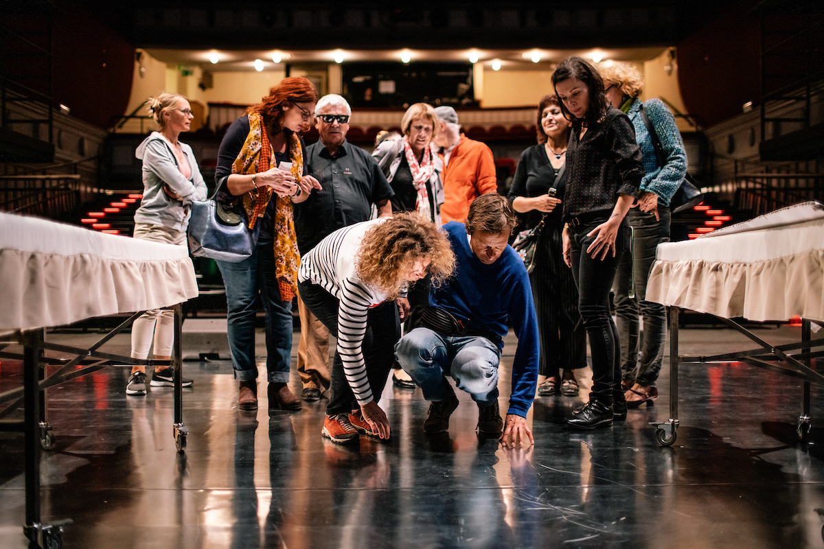 Une dizaine de personnes sur la scène d’un théâtre, les gradins à l’arrière-plan. Au premier plan, une personne accroupie touche du bout des doigts le revêtement noir de la scène.