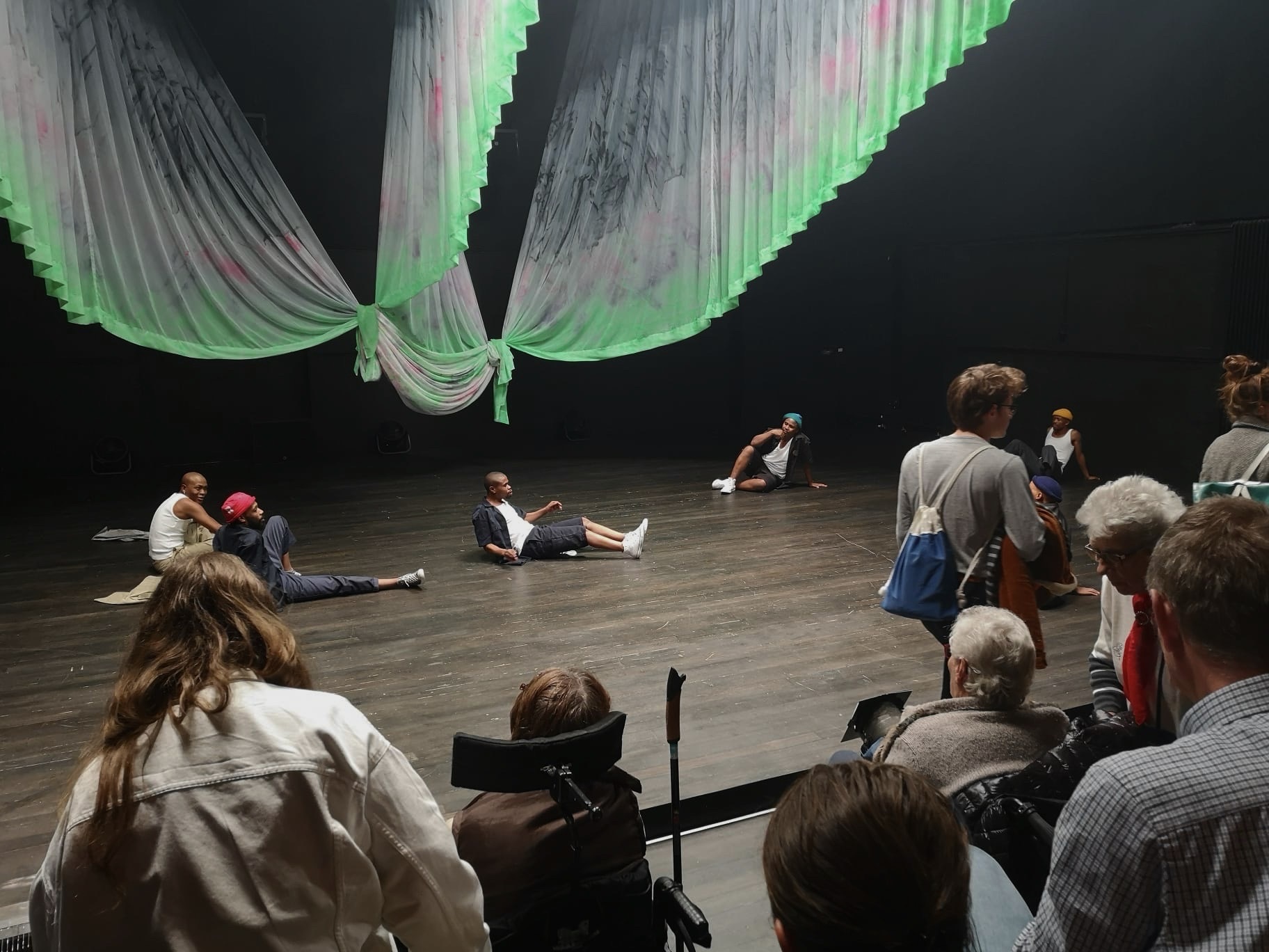 Un public d’âge varié, assis, debout ou en train de se déplacer dans les gradins. Sur la scène, six jeunes hommes, confortablement assis au sol, se regardent. Des bandes de tissu pendent du plafond comme des ailes, l’éclairage de la scène est uniforme.