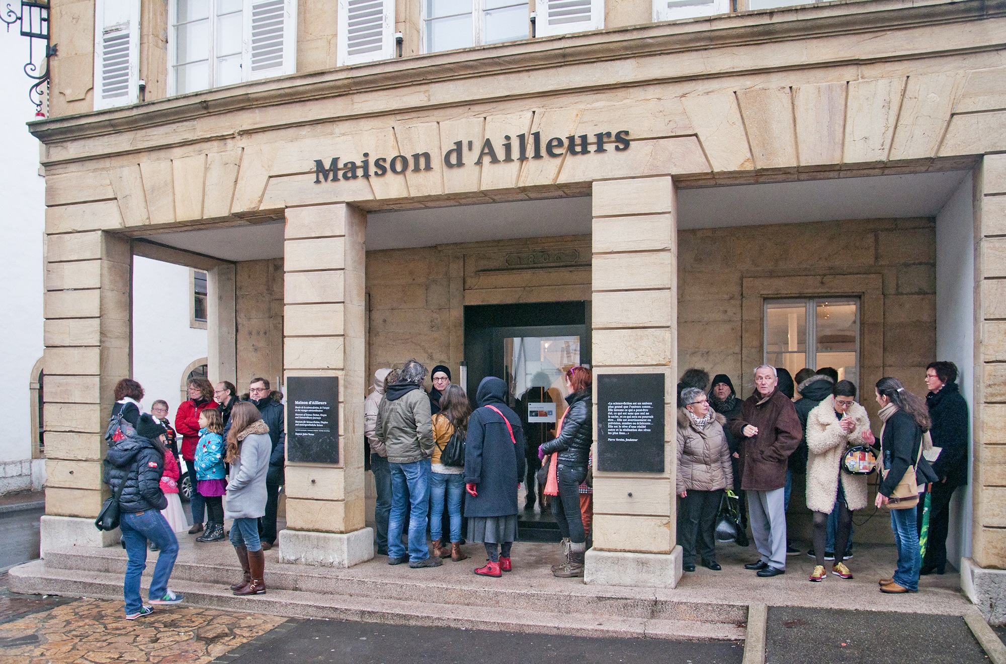 Des enfants, des jeunes et des moins jeunes se tiennent sous le porche d’un bâtiment. Le porche est accessible par deux marches ou par une rampe goudronnée sur le côté droit. L’inscription « Maison d’Ailleurs » figure au milieu de la façade.