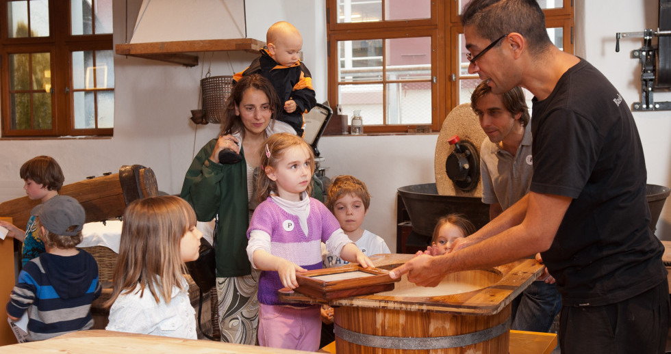 Vier Kinder und drei Erwachsene stehen um einen Holzbottich mit Wasser. Das Mädchen im Zentrum hält einen Papier-Schöpfrahmen und schaut fragend die männliche Vermittlungsperson an. Im Hintergrund sieht man andere Utensilien zur Papierherstellung.