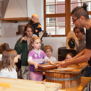 Vier Kinder und drei Erwachsene stehen um einen Holzbottich mit Wasser. Das Mädchen im Zentrum hält einen Papier-Schöpfrahmen und schaut fragend die männliche Vermittlungsperson an. Im Hintergrund sieht man andere Utensilien zur Papierherstellung.