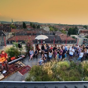 Auf einer begrünten Dachterrasse verfolgen über vierzig Menschen unterschiedlichen Alters ein Konzert eines Musikers mit Gitarre. Er steht auf einer Bühne aus Bahnpaletten zwischen zwei Keybords und einer Elektrogitarre sowie Mischpult und zwei Boxen.